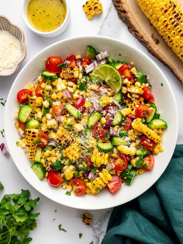 Overhead image of finished corn salad in a large white serving bowl on a white marble surface. The salad is made with charred corn, chopped cucumber, halved cherry tomatoes, diced red onion, and chopped cilantro, then topped with a sprinkle of Cotija cheese and garnished with a lime wedge. Around the bowl are a small white ramekin of lime dressing, a dish of Cotija cheese, fresh cilantro, lime wedges, and a grilled ear of corn resting on a round wooden board. A dark teal cloth napkin is tucked beside the bowl.