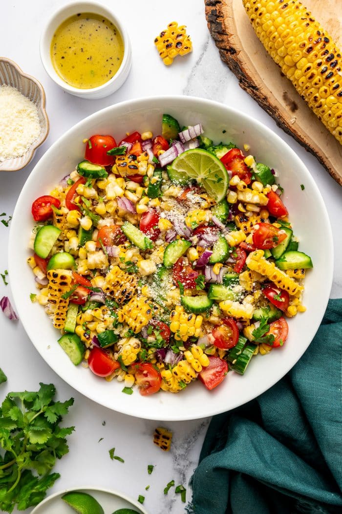Overhead image of finished corn salad in a large white serving bowl on a white marble surface. The salad is made with charred corn, chopped cucumber, halved cherry tomatoes, diced red onion, and chopped cilantro, then topped with a sprinkle of Cotija cheese and garnished with a lime wedge. Around the bowl are a small white ramekin of lime dressing, a dish of Cotija cheese, fresh cilantro, lime wedges, and a grilled ear of corn resting on a round wooden board. A dark teal cloth napkin is tucked beside the bowl.