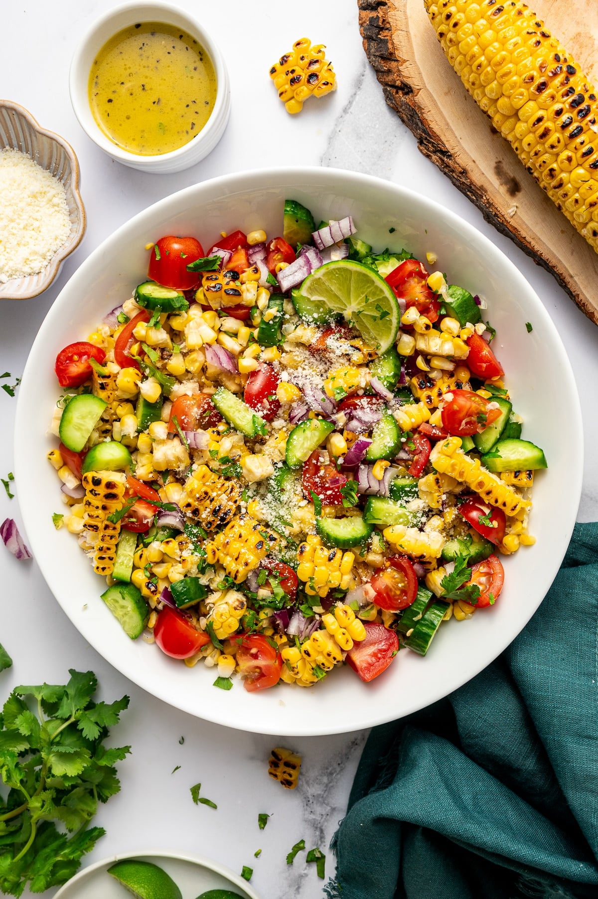 Overhead image of finished grilled corn salad in a large white serving bowl on a white marble surface. The salad is made with charred corn, chopped cucumber, halved cherry tomatoes, diced red onion, and chopped cilantro, then topped with a sprinkle of Cotija cheese and garnished with a lime wedge. Around the bowl are a small white ramekin of lime dressing, a dish of Cotija cheese, fresh cilantro, lime wedges, and a grilled ear of corn resting on a round wooden board. A dark teal cloth napkin is tucked beside the bowl.