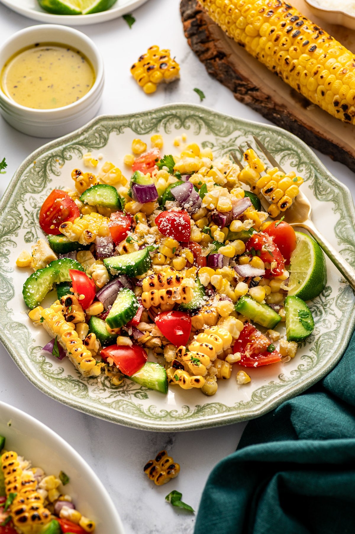 Overhead image of a serving of corn salad plated on a cream-colored dish with a green decorative border. The salad includes charred corn, chopped cucumber, cherry tomatoes, diced red onion, cilantro, and a light sprinkle of Cotija cheese, with a lime wedge on the side. In the background are a small bowl of dressing, a grilled ear of corn on a wooden board, and a dark teal cloth napkin on the white marble surface.