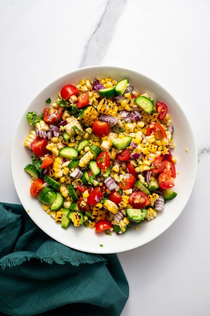 Overhead image of mixed corn salad in a large white bowl on a white marble surface. The salad includes charred corn kernels, halved cherry tomatoes, chopped cucumber, diced red onion, and flecks of fresh cilantro. A dark teal cloth napkin is placed beside the bowl.
