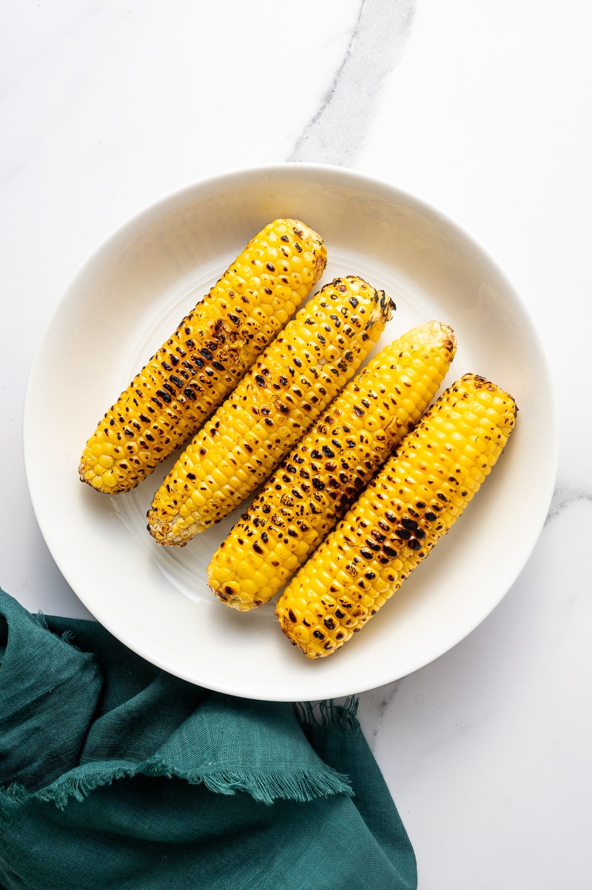 Overhead image of four ears of grilled corn with light char marks resting on a large round white plate on a white marble surface. A dark teal cloth napkin is draped in the lower left corner.