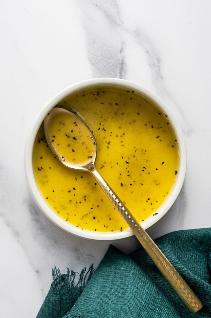 Close overhead image of bright yellow lime dressing in a small white bowl with visible black pepper specks. A gold spoon rests inside the bowl, and a dark teal cloth napkin lies to the left on the white marble surface.