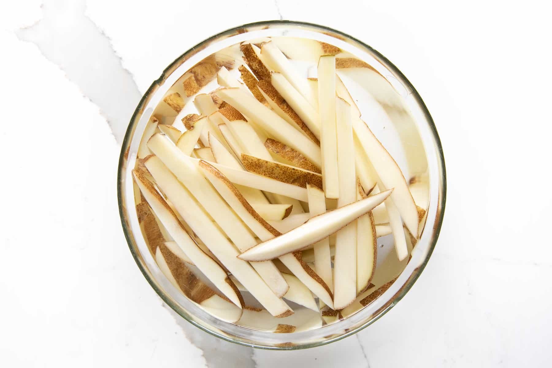 Raw potato sticks soaking in a glass bowl of cold water to remove excess starch before air frying.