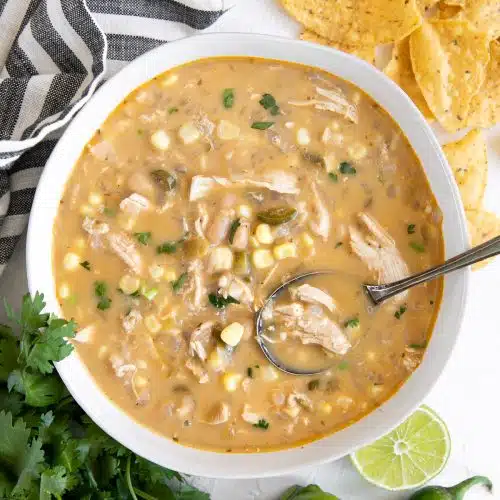 White shallow bowl filled with creamy white chicken chili garnished with crushed corn tortilla chips and fresh cilantro on a table with a white and navy striped napkin and whole tortilla chips.