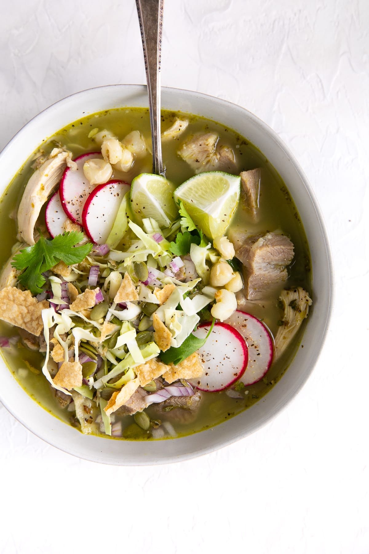 White bowl filled with pozole verde garnished with shredded cabbage, tortilla chops, cilantro, sliced radish, and lime juice.
