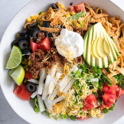 Large white bowl filled with a bed of fresh greens topped with ground beef taco meat, shredded cheese, avocado, olives, tomatoes, and sour cream.