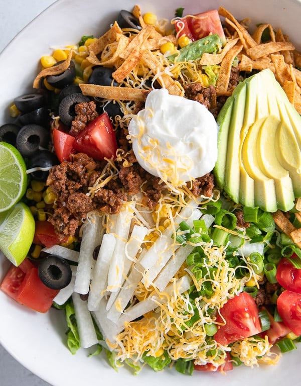 Large white bowl filled with a bed of fresh greens topped with ground beef taco meat, shredded cheese, avocado, olives, tomatoes, and sour cream.