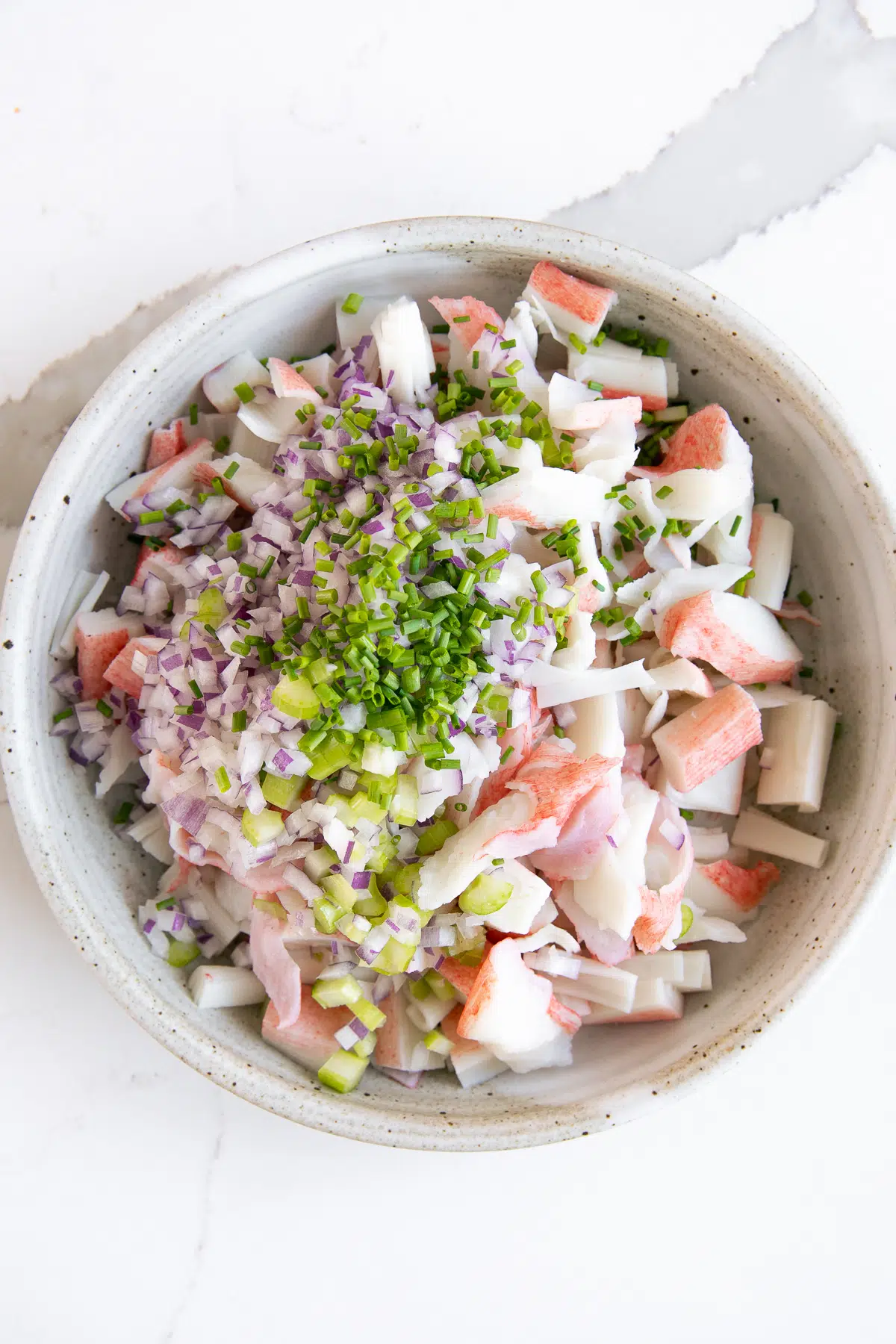 Large white bowl filled with imitation crab, diced red onion, chopped celery, and chopped chives.