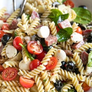 Large white salad bowl filled with a cold Italian pasta salad made with cherry tomatoes, fresh spinach, mozzarella pearls, salami, black olives, artichoke hearts and rotini pasta all tossed in a homemade Italian dressing.