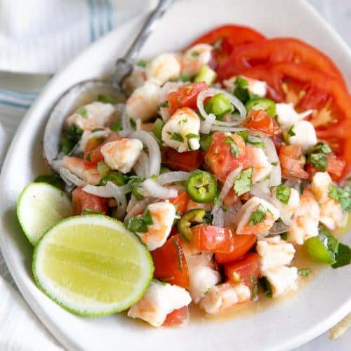 White shallow serving plate filled with shrimp ceviche made with chopped shrimp, sliced onion, chopped bell peppers, and tomatoes in citrus juice with chopped cilantro and chopped jalapenos.