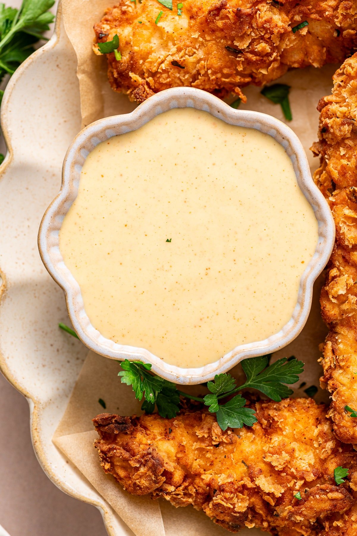 A small bowl of rich, golden honey mustard sauce sits on a larger white plate, surrounded by perfectly crisp, golden-brown chicken tenders—ready for dipping.