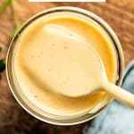 Overhead view of glass jar filled with creamy honey mustard sauce. A spoon is raised slightly out of the sauce, showcasing its thick, dip-ready texture.