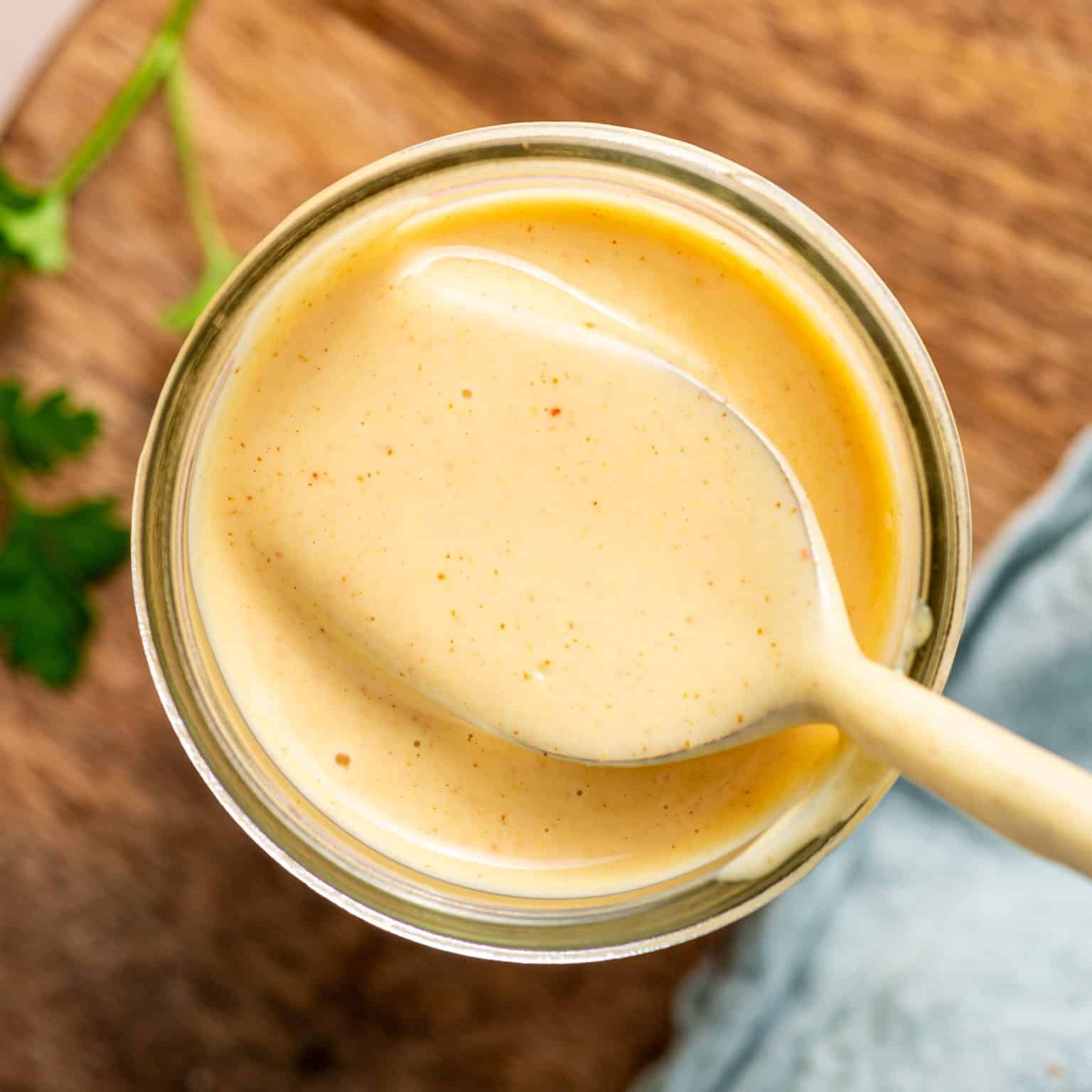 Overhead view of glass jar filled with creamy honey mustard sauce. A spoon is raised slightly out of the sauce, showcasing its thick, dip-ready texture.