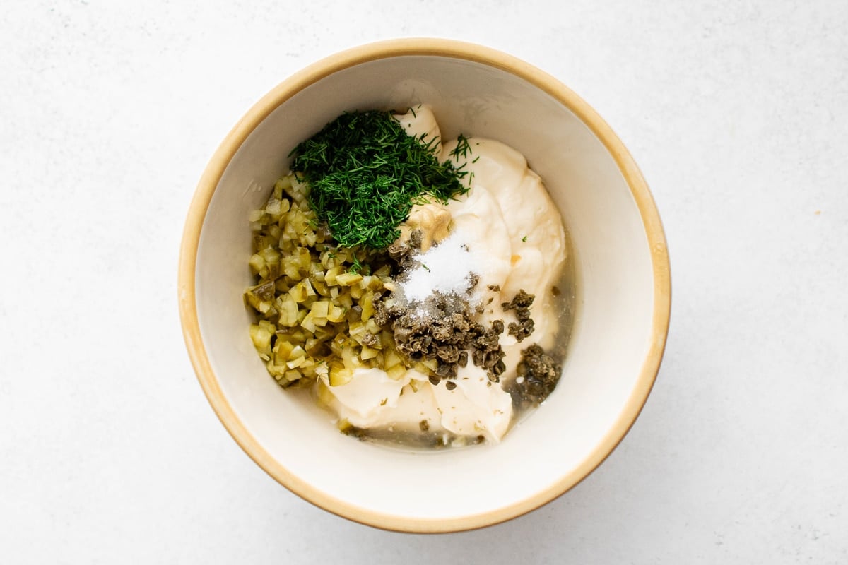 Overhead view of a mixing bowl filled with mayonnaise, chopped dill pickles, minced fresh dill, chopped capers, Dijon mustard, sugar, and lemon juice before mixing.