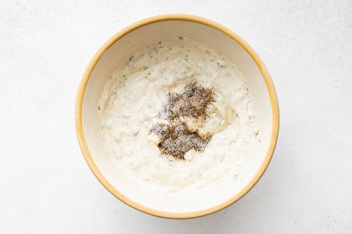 Overhead view of partially mixed tartar sauce in a bowl with black pepper and salt sitting on top, ready to be stirred in.