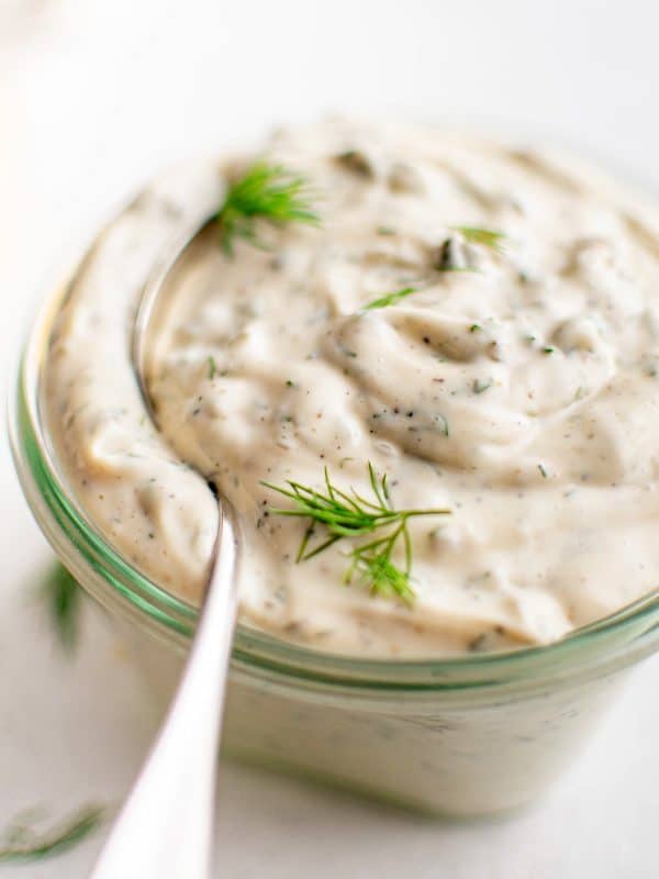Close-up of tartar sauce in a small glass jar with a spoon dipped in, topped with fresh dill.