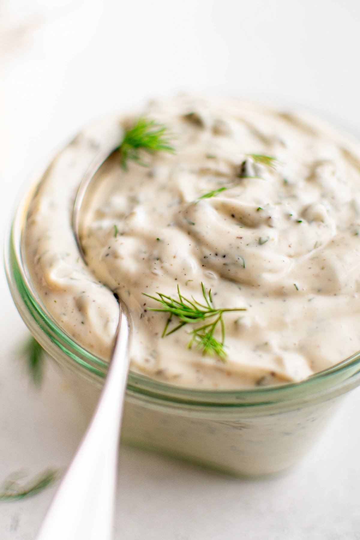 Close-up of tartar sauce in a small glass jar with a spoon dipped in, topped with fresh dill.