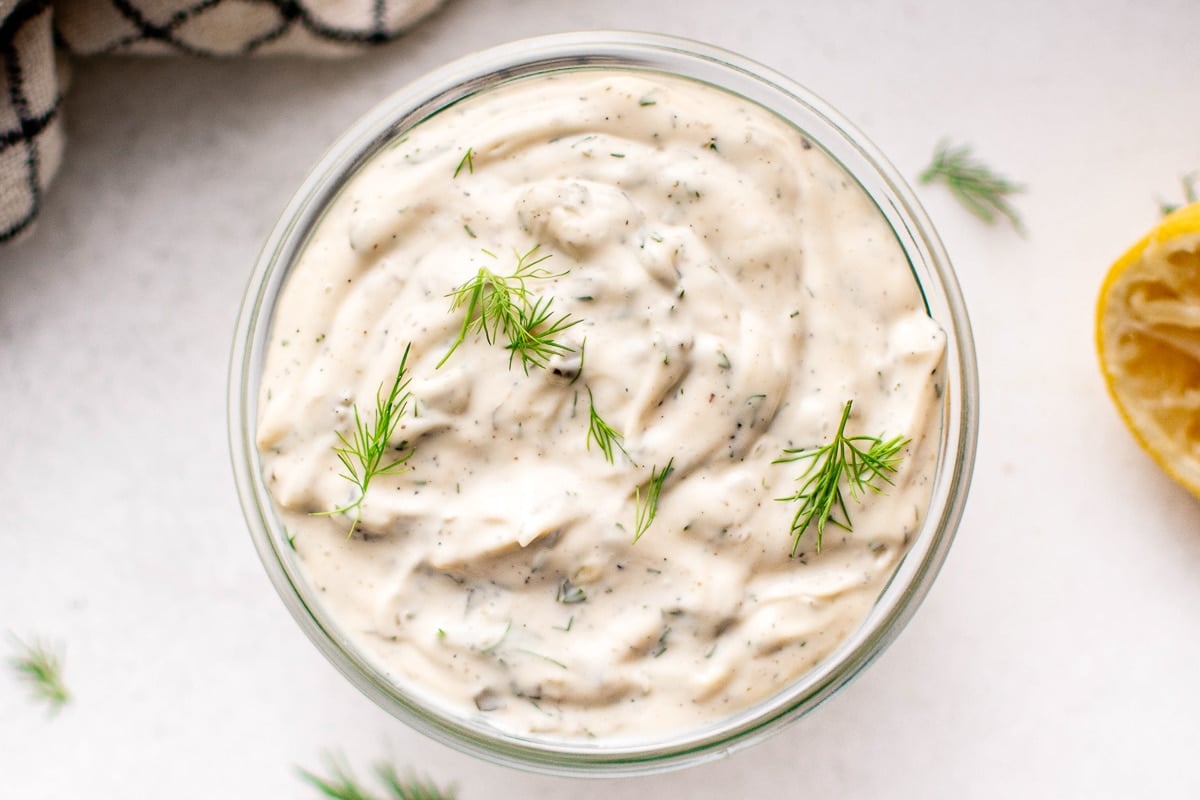 Overhead view of a glass jar filled with tartar sauce, garnished with dill sprigs, and surrounded by fresh herbs and a squeezed lemon half.