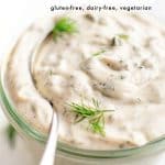 Close-up of homemade tartar sauce in a glass jar, garnished with fresh dill and labeled as gluten-free, dairy-free, and vegetarian. A spoon is dipped into the creamy sauce.