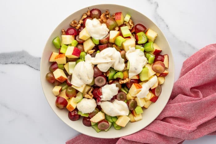 Overhead view of a light beige bowl filled with chopped apples, halved red grapes, celery, and walnuts on a white marble surface, topped with dollops of creamy mayonnaise dressing, with a pink cloth napkin draped along the lower right side.
