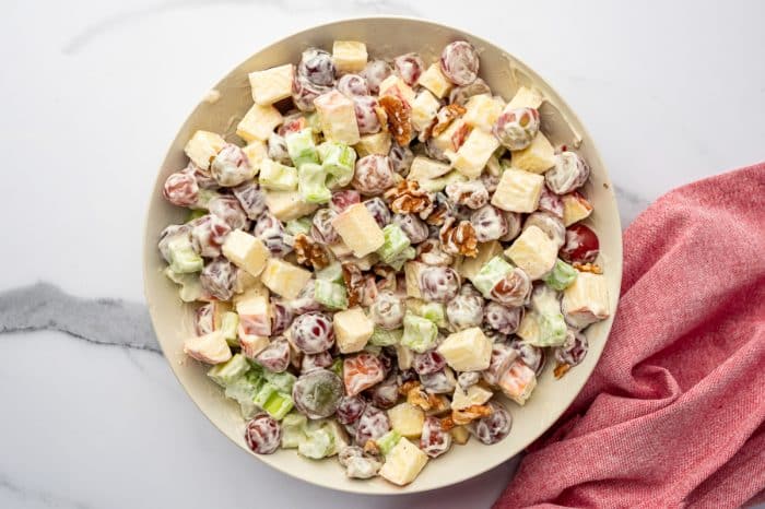 Overhead view of a light beige bowl filled with mixed Waldorf salad on a white marble surface, with chopped apples, halved red grapes, celery, and walnuts coated in a creamy mayonnaise dressing, and a pink cloth napkin draped along the lower right side.