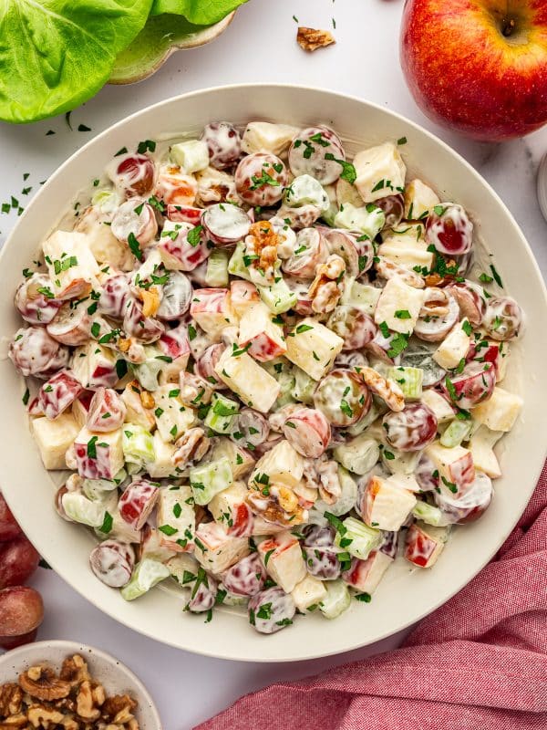 Overhead close view of a light beige bowl filled with creamy Waldorf salad on a white marble surface, made with chopped red-skinned apples, halved red grapes, celery, and walnuts, garnished with chopped green parsley, with butter lettuce in a bowl at the upper left, a whole red apple at the upper right, a small bowl of walnuts at the lower left, loose grapes around the edges, and a pink cloth napkin draped along the lower right side.