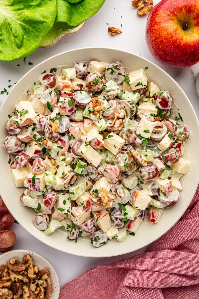 Overhead close view of a light beige bowl filled with creamy Waldorf salad on a white marble surface, made with chopped red-skinned apples, halved red grapes, celery, and walnuts, garnished with chopped green parsley, with butter lettuce in a bowl at the upper left, a whole red apple at the upper right, a small bowl of walnuts at the lower left, loose grapes around the edges, and a pink cloth napkin draped along the lower right side.