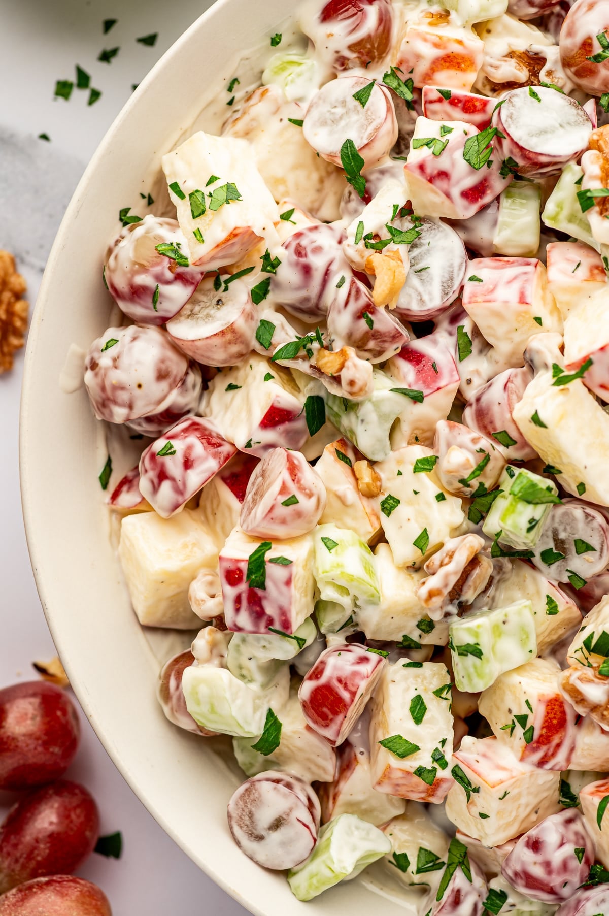Close-up of creamy Waldorf salad in a light beige bowl, showing chopped red-skinned apples, halved red grapes, celery, and walnuts coated in dressing and garnished with chopped green parsley, with a few loose grapes and walnut pieces blurred around the edges.