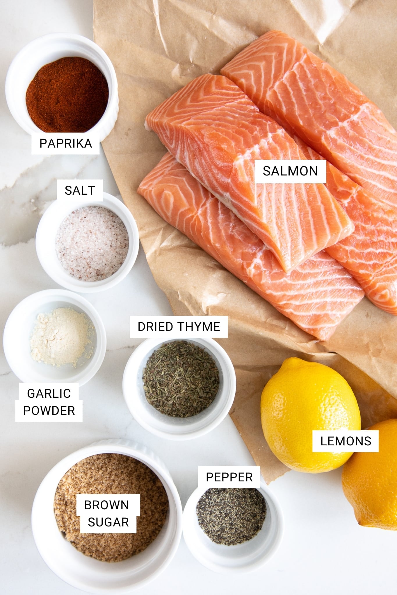 Overhead image of raw salmon fillets on brown butcher paper surrounded by white ramekins filled with paprika, salt, garlic powder, dried thyme, brown sugar, and black pepper on a white marble surface, with whole lemons on the side. Each ingredient is labeled with black text on white text overlays, including salmon, paprika, salt, garlic powder, dried thyme, brown sugar, pepper, and lemons.