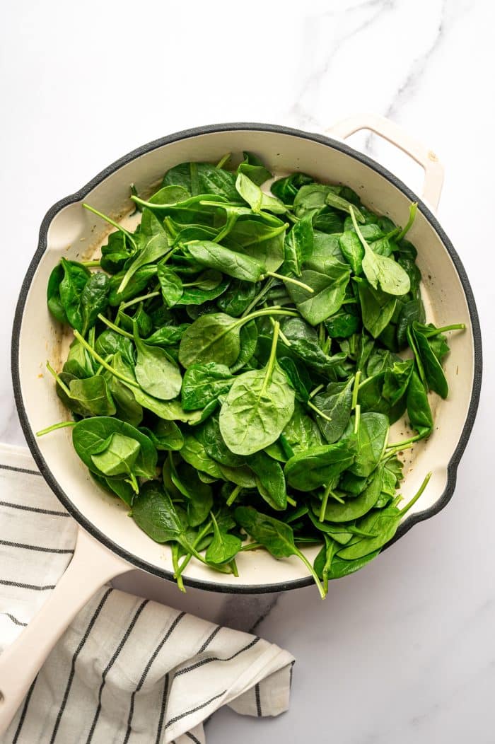 A large cream-colored skillet is filled with fresh baby spinach piled high over the cream sauce base. The bright green spinach leaves sit on top of the pale sauce, and the skillet rests on a white marble surface with a cream-colored kitchen towel with thin black stripes nearby.