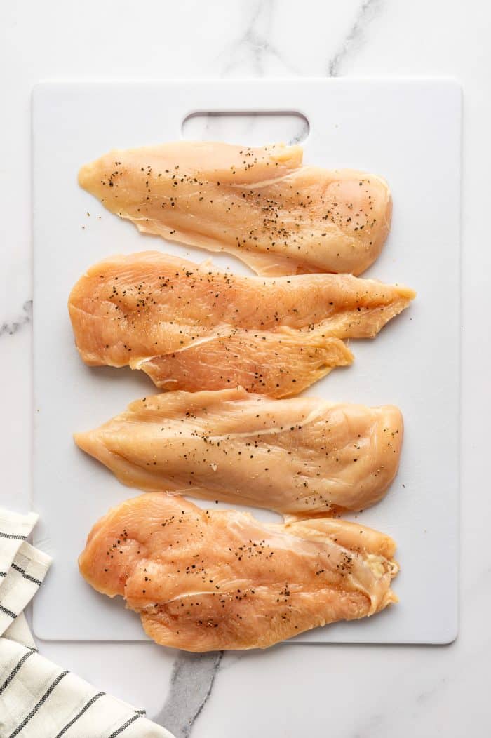Four raw boneless skinless chicken breast cutlets seasoned with black pepper are arranged vertically on a white plastic cutting board with a handle cutout, set on a white marble surface. A cream-colored kitchen towel with thin black stripes is partially visible in the bottom left corner.