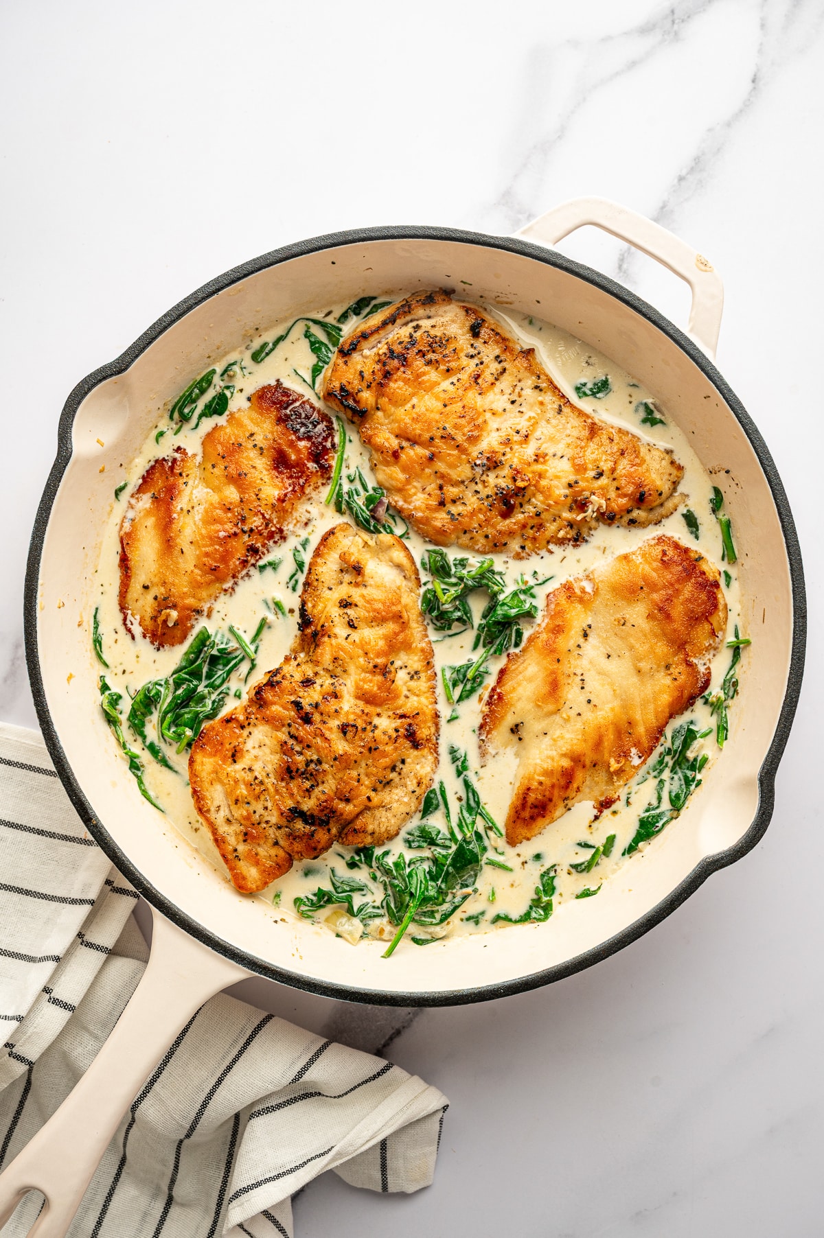 Four golden brown chicken cutlets are nestled back into a pale cream sauce with wilted spinach in a large cream-colored skillet. The skillet is set on a white marble surface with a cream-colored kitchen towel with thin black stripes at the lower left.