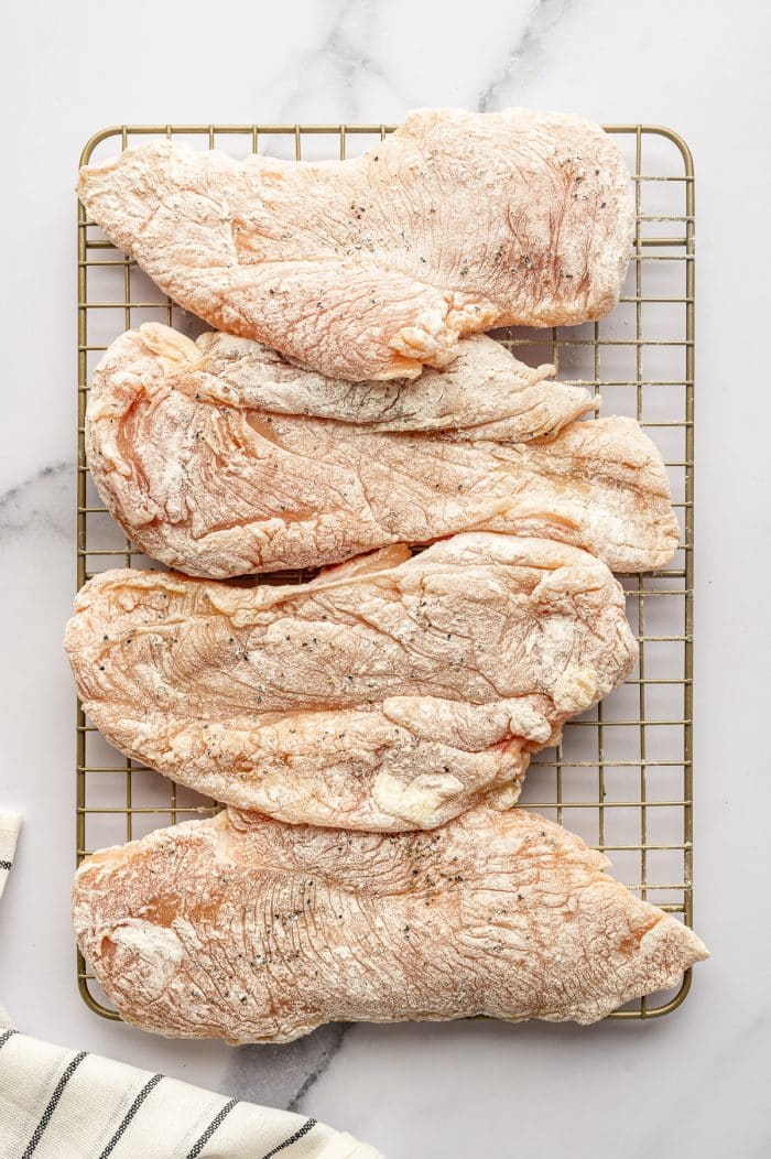 Four raw chicken cutlets lightly dredged in flour are arranged on a gold wire rack set over a white marble surface. The flour coating is visible across the tops of the chicken, and a cream-colored kitchen towel with thin black stripes appears in the lower left corner.