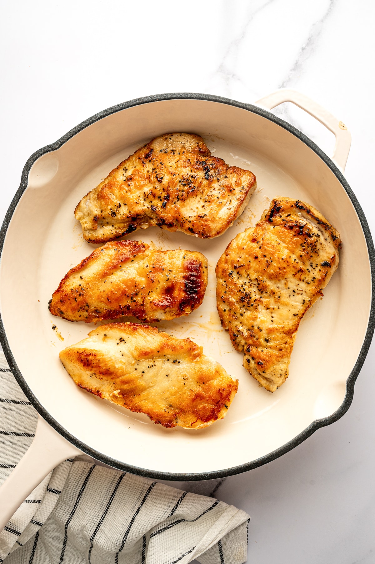 Four golden brown chicken cutlets are seared in a large round cream-colored skillet with a light interior and side handle, set on a white marble surface. The chicken has browned edges and black pepper seasoning visible on top. A cream-colored kitchen towel with thin black stripes is tucked along the lower left side.
