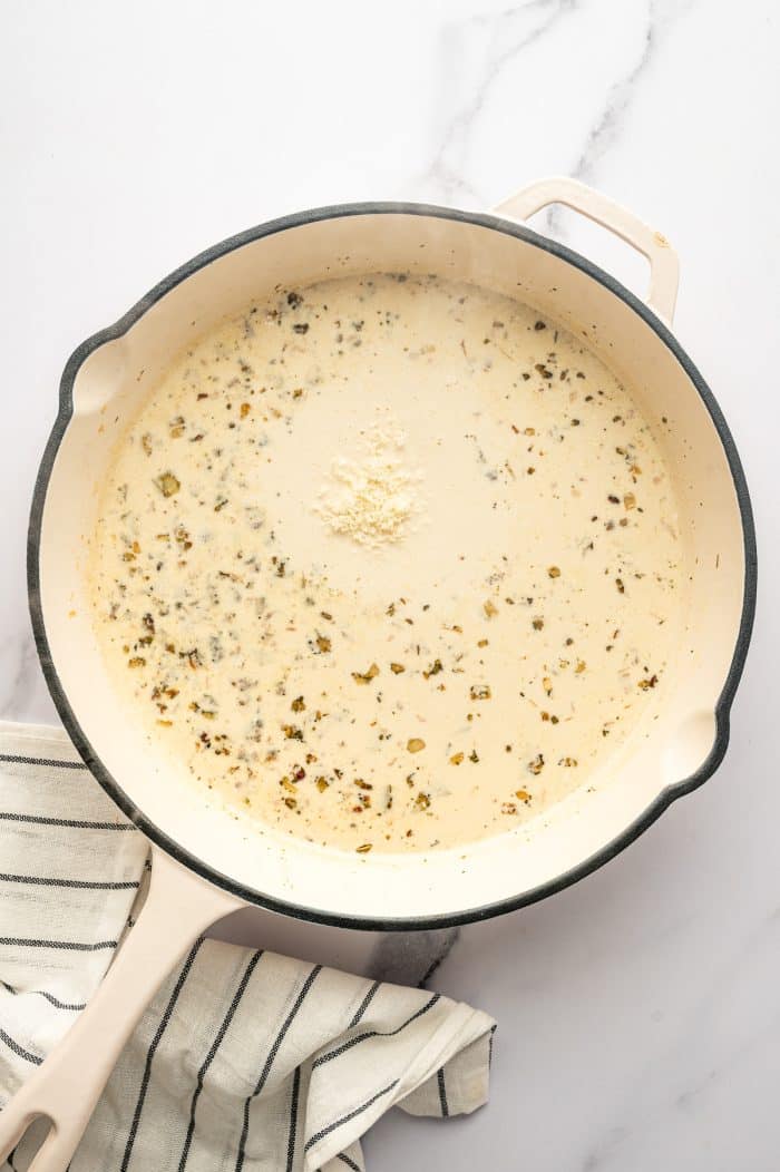 A large cream-colored skillet contains a pale off-white cream sauce speckled with cooked shallots and seasoning, with a small mound of grated parmesan cheese in the center. The skillet sits on a white marble surface beside a cream-colored kitchen towel with thin black stripes.