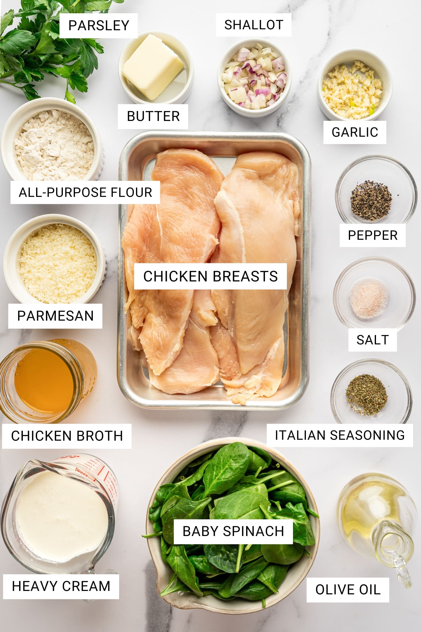 Overhead ingredient photo for Chicken Florentine on a white marble surface. Raw chicken breasts sit in a silver metal pan in the center and are labeled “CHICKEN BREASTS.” Surrounding ingredients include fresh parsley labeled “PARSLEY,” butter labeled “BUTTER,” chopped shallot labeled “SHALLOT,” minced garlic labeled “GARLIC,” black pepper labeled “PEPPER,” salt labeled “SALT,” dried Italian seasoning labeled “ITALIAN SEASONING,” olive oil labeled “OLIVE OIL,” baby spinach labeled “BABY SPINACH,” heavy cream labeled “HEAVY CREAM,” chicken broth labeled “CHICKEN BROTH,” parmesan labeled “PARMESAN,” and all-purpose flour labeled “ALL-PURPOSE FLOUR.” All labels appear in black uppercase text inside white rectangular boxes.