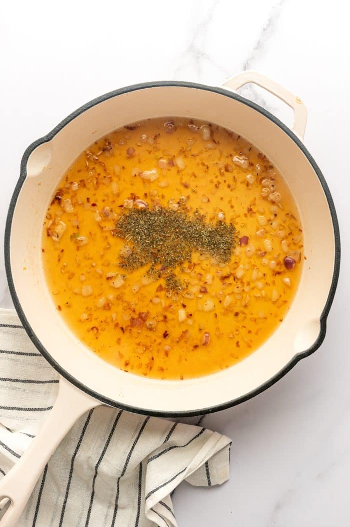 A large cream-colored skillet holds chopped shallots simmering in golden chicken broth with a mound of dried Italian seasoning in the center. The skillet sits on a white marble surface with a cream-colored kitchen towel with thin black stripes along the lower left side.