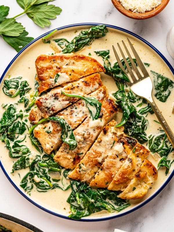 Overhead view of sliced Chicken Florentine served in a shallow cream-colored bowl with a dark blue rim, filled with creamy spinach sauce. One chicken cutlet has been sliced into strips and arranged in the center of the bowl, topped with sauce and wilted baby spinach. A gold fork rests on the right side of the bowl. Fresh parsley leaves are scattered on the white marble background, and a small wooden bowl of grated parmesan is visible in the top right corner.