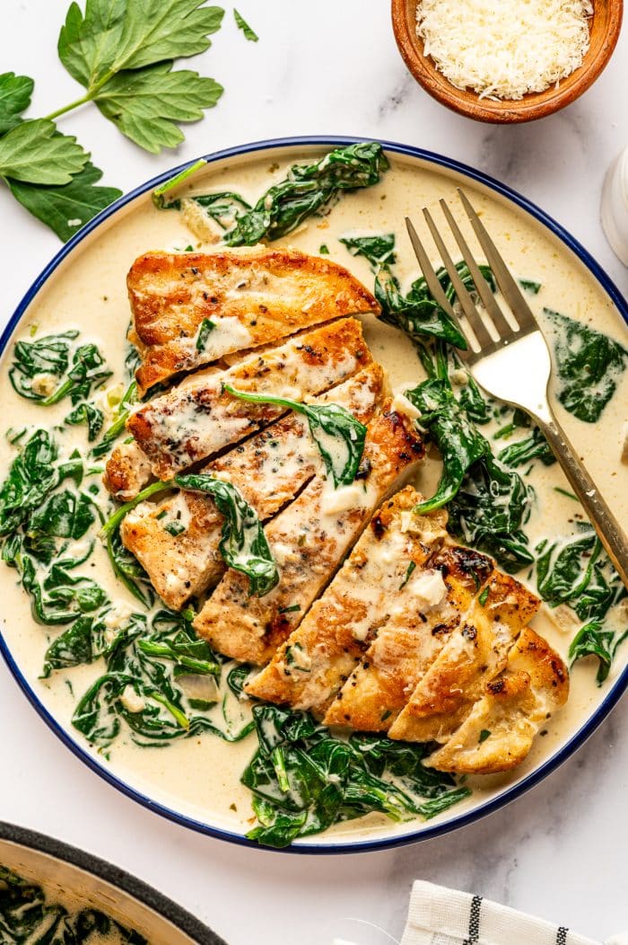 Overhead view of sliced Chicken Florentine served in a shallow cream-colored bowl with a dark blue rim, filled with creamy spinach sauce. One chicken cutlet has been sliced into strips and arranged in the center of the bowl, topped with sauce and wilted baby spinach. A gold fork rests on the right side of the bowl. Fresh parsley leaves are scattered on the white marble background, and a small wooden bowl of grated parmesan is visible in the top right corner.