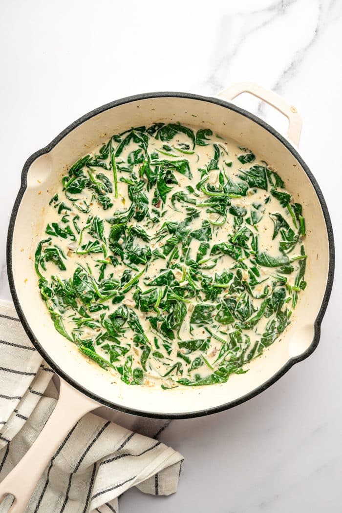 A large cream-colored skillet holds a pale parmesan cream sauce with wilted dark green spinach evenly distributed throughout. The skillet sits on a white marble surface with a cream-colored kitchen towel with thin black stripes at the lower left.