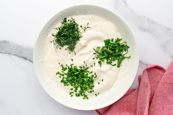 Overhead view of a large white bowl filled with creamy ranch base, topped with three piles of fresh herbs: chopped dill at the upper left, chopped parsley at the upper right, and chopped chives in the center, on a white marble surface with a pink kitchen towel to the right.