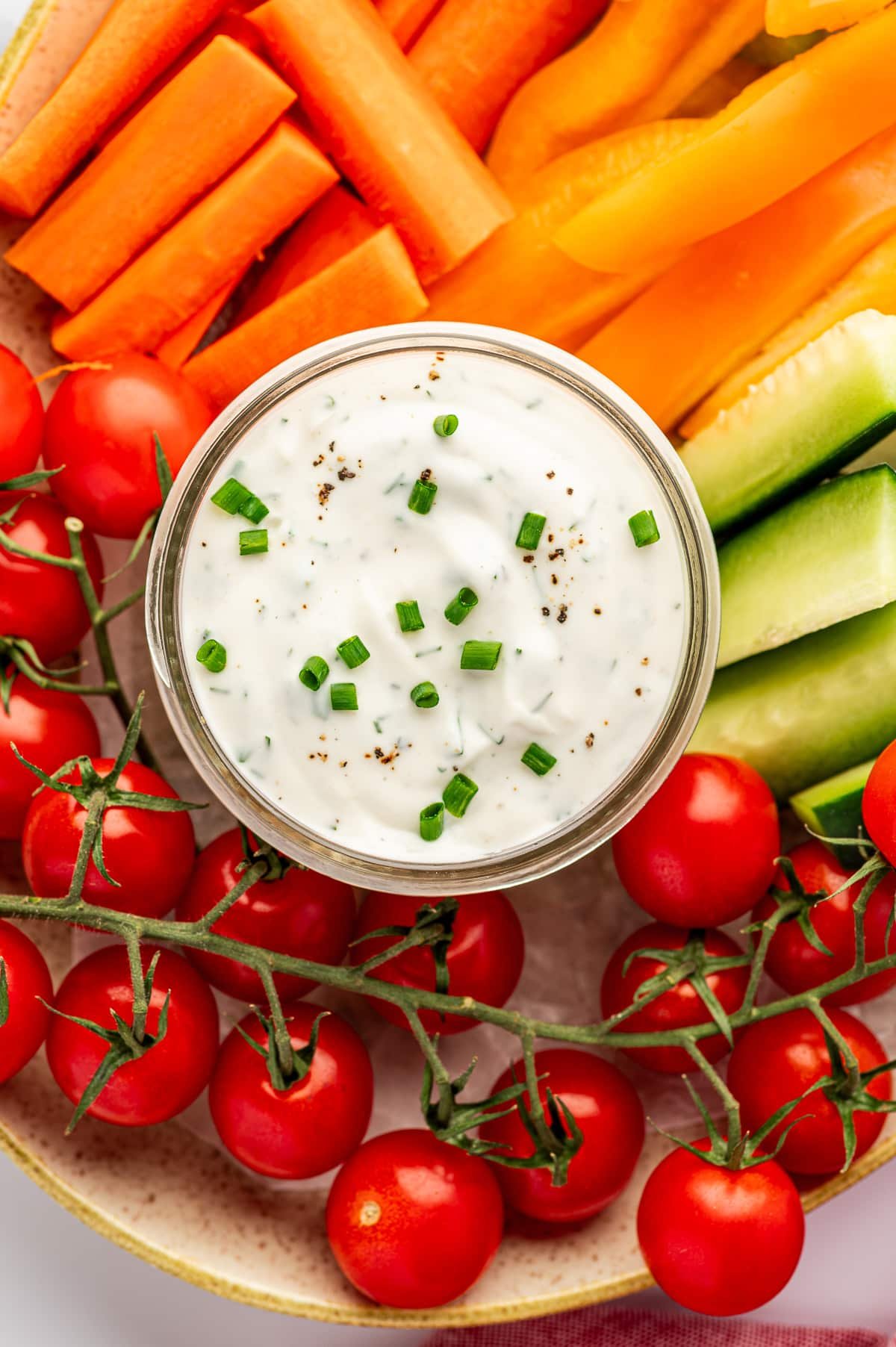 Close-up overhead view of a small glass jar of ranch dressing topped with chopped chives and black pepper, surrounded by bright orange carrot sticks, orange bell pepper slices, green cucumber spears, and red cherry tomatoes on the vine on a speckled beige platter.
