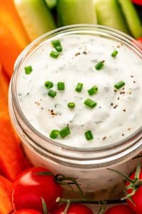 Tight close-up of the top of a clear glass jar filled with creamy ranch dressing, sprinkled with chopped chives and black pepper, with blurred carrot sticks, cucumber spears, and cherry tomatoes in the background.