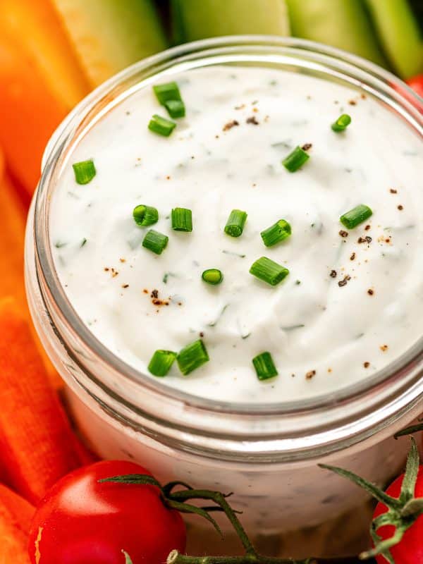 Tight close-up of the top of a clear glass jar filled with creamy ranch dressing, sprinkled with chopped chives and black pepper, with blurred carrot sticks, cucumber spears, and cherry tomatoes in the background.