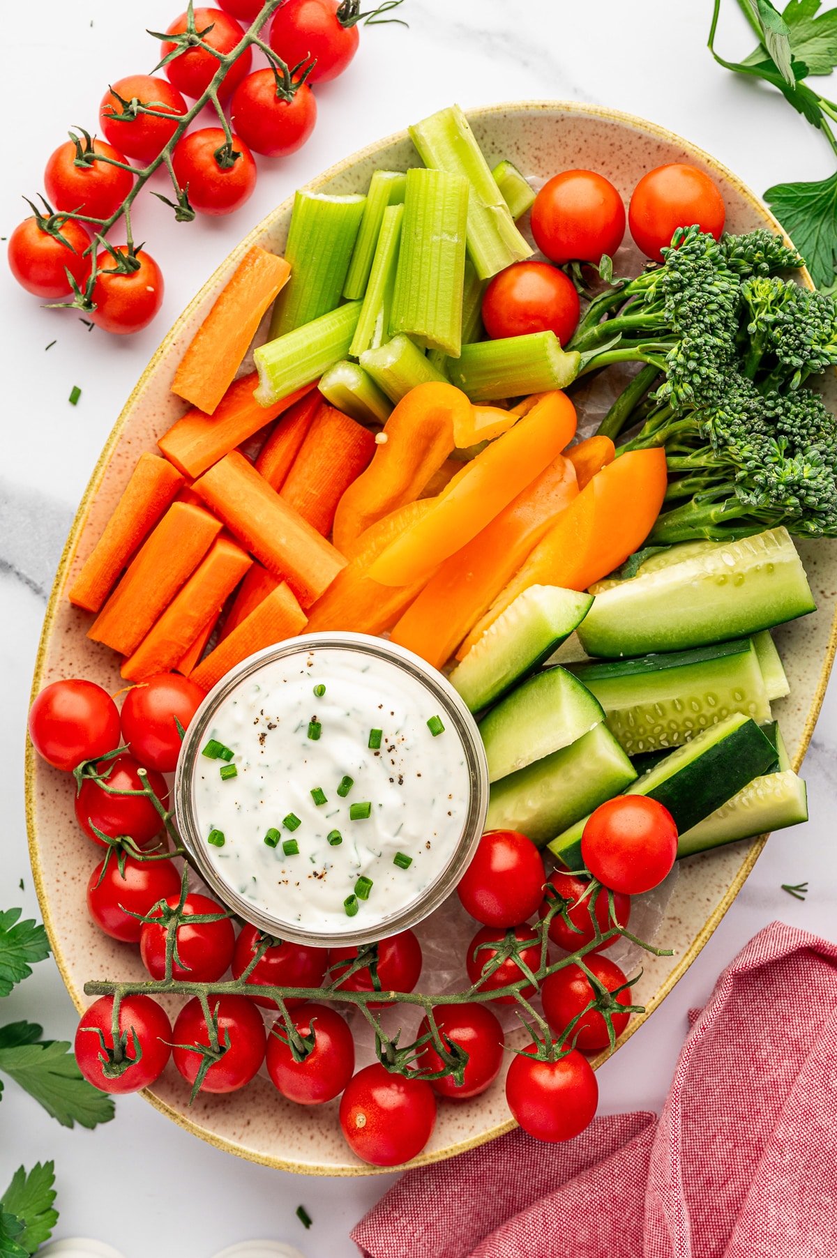 Vertical overhead shot of a speckled beige oval platter filled with fresh vegetables including carrot sticks, celery sticks, orange bell pepper slices, cucumber spears, broccolini, and cherry tomatoes on the vine, with a small glass jar of ranch dressing in the center topped with chopped chives and black pepper, and a pink kitchen towel at the lower right.