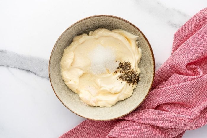 Overhead view of a small beige speckled bowl filled with mayonnaise on a white marble surface, topped with white sugar, salt, and cracked black pepper, with a pink cloth napkin draped along the lower right side.