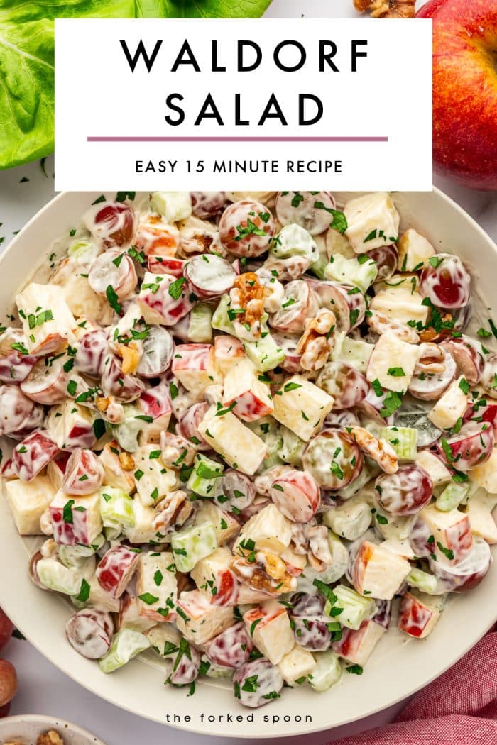 Vertical Pinterest graphic showing a light beige bowl filled with creamy Waldorf salad on a pale background, made with chopped red-skinned apples, halved red grapes, celery, and walnuts, garnished with chopped green parsley. Bright green lettuce leaves appear in the upper left corner, and a whole red apple is visible in the upper right corner. A large light gray text box overlays the upper center of the image with black text reading “WALDORF SALAD” and smaller black text below reading “EASY 15 MINUTE RECIPE,” separated by a muted pink line. Small black text at the bottom reads “the forked spoon.”