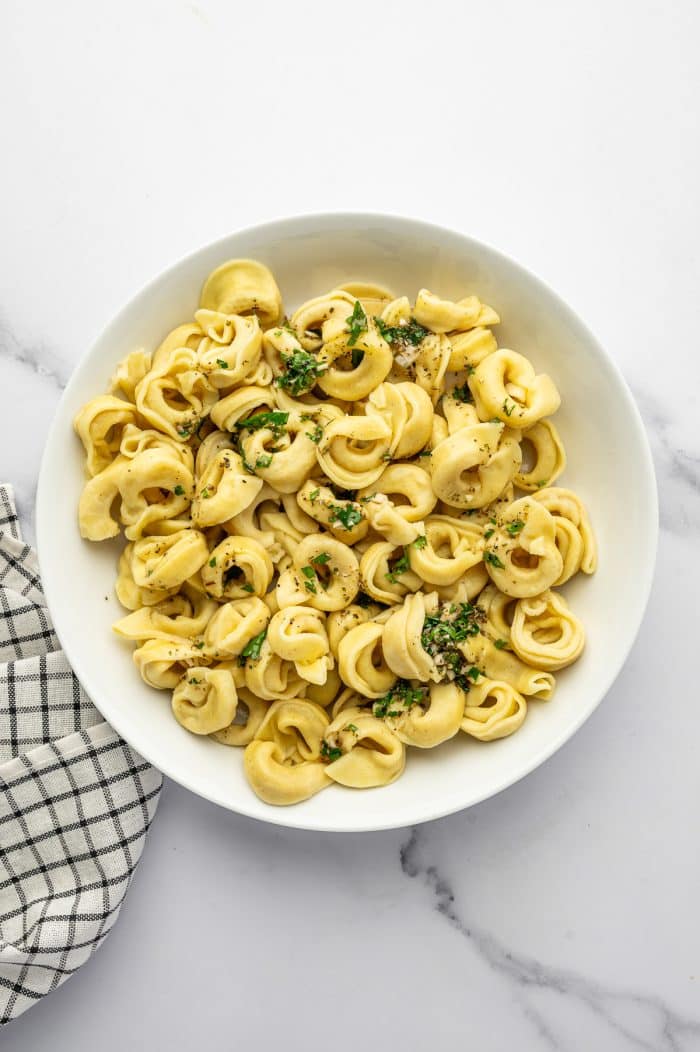 Overhead image of cooked cheese tortellini in a large white bowl tossed with homemade Italian dressing, chopped parsley, and dried herbs. The bowl sits on a white marble surface with a black and white checkered towel along the left side.