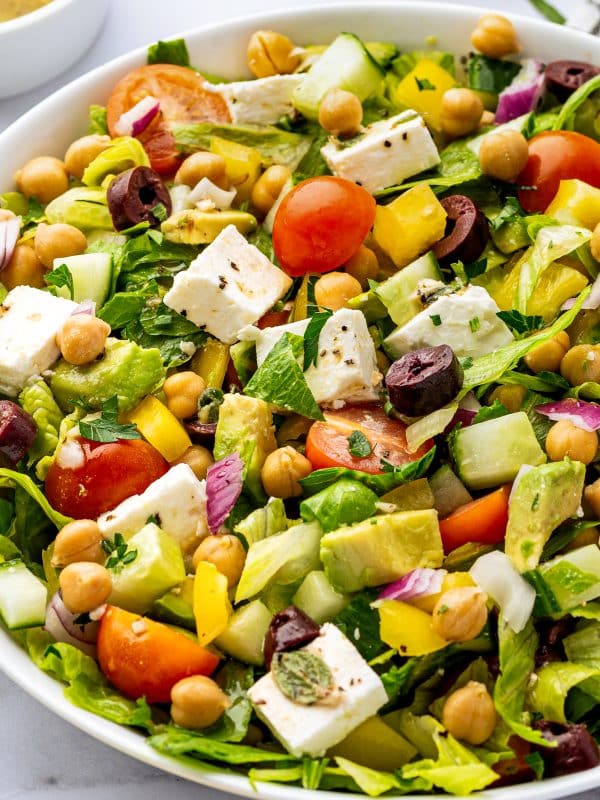 Close-up side view of Mediterranean chopped salad with romaine lettuce, chickpeas, cubed feta cheese, avocado, cucumber, cherry tomatoes, yellow bell pepper, red onion, Kalamata olives, parsley, and black pepper in a white bowl.